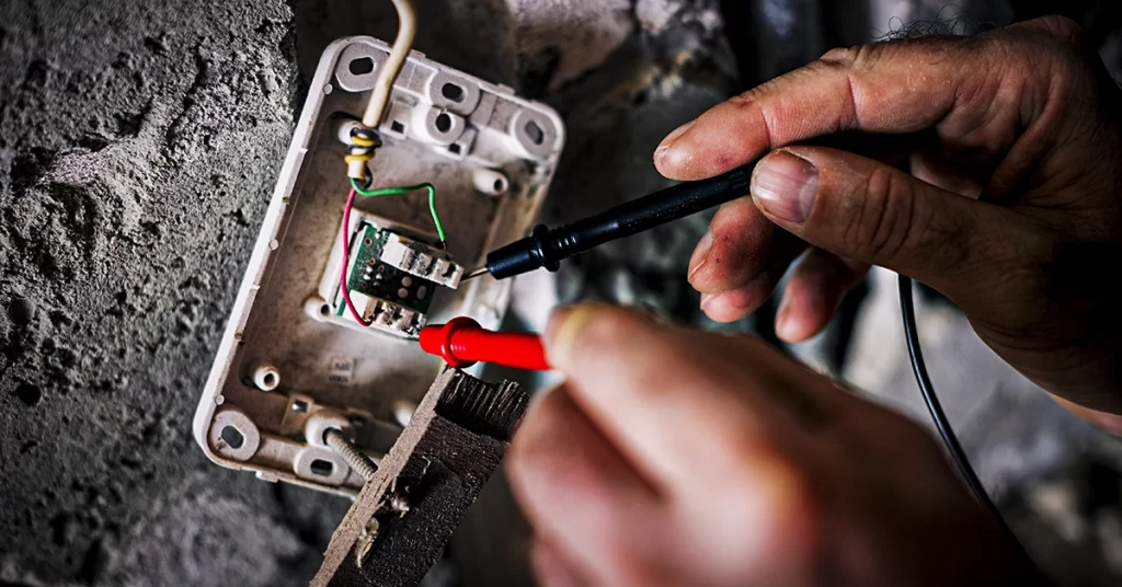 Mãos testando instalação elétrica com multímetro em parede, representando verificação de risco de incêndio