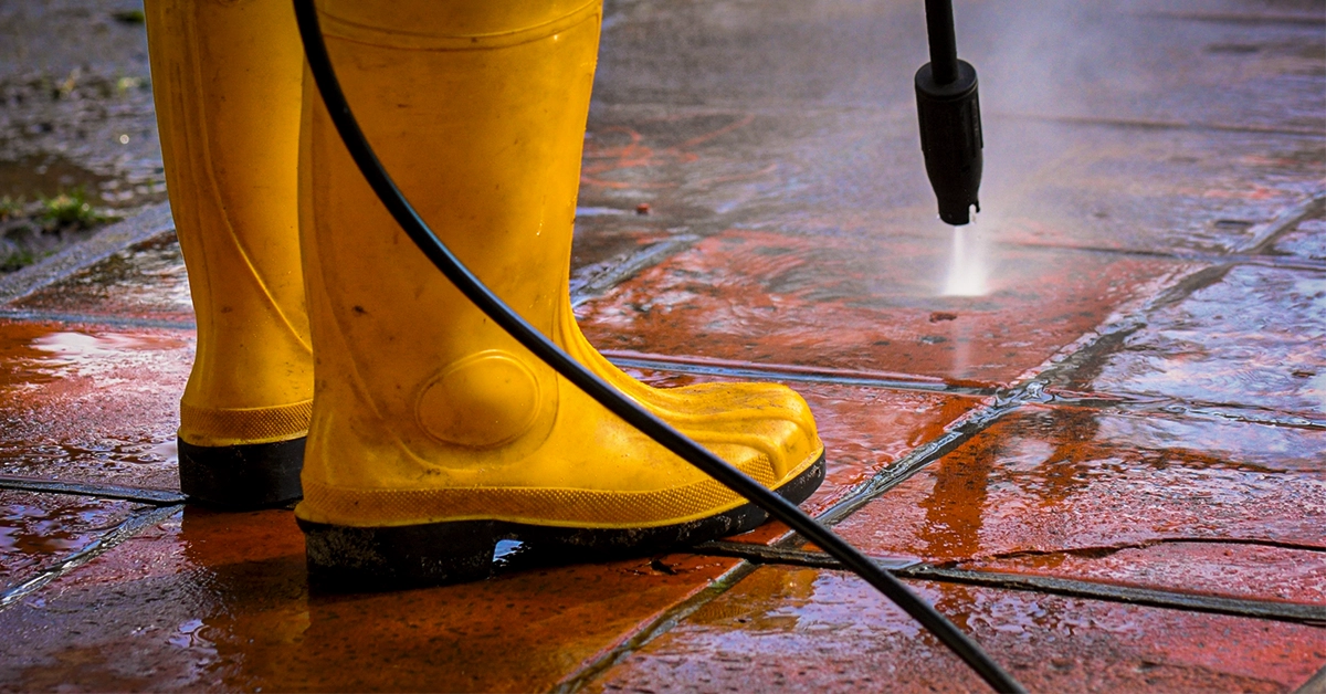Capa do artigo Quedas em Pisos Escorregadios no Terreiro: O que Fazer para Evitar Acidentes, com uma pessoa usando botas de borracha amarelas para lavar um quintal com um jato de água pressurizada